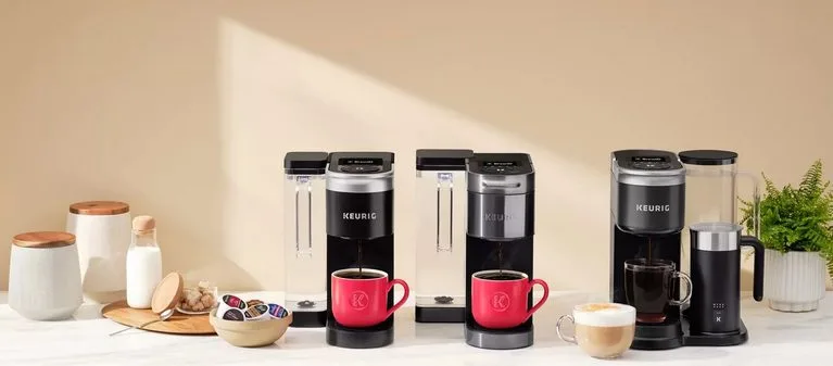Trzy ekspresy do kawy Keurig przygotowujące kawę w czerwonych i czarnych filiżankach, obok spieniacza mleka
