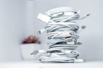 Zwijane białe kable USB i Lightning ułożone w pionowy stos na jasnym tle, w tle rozmyta roślina.